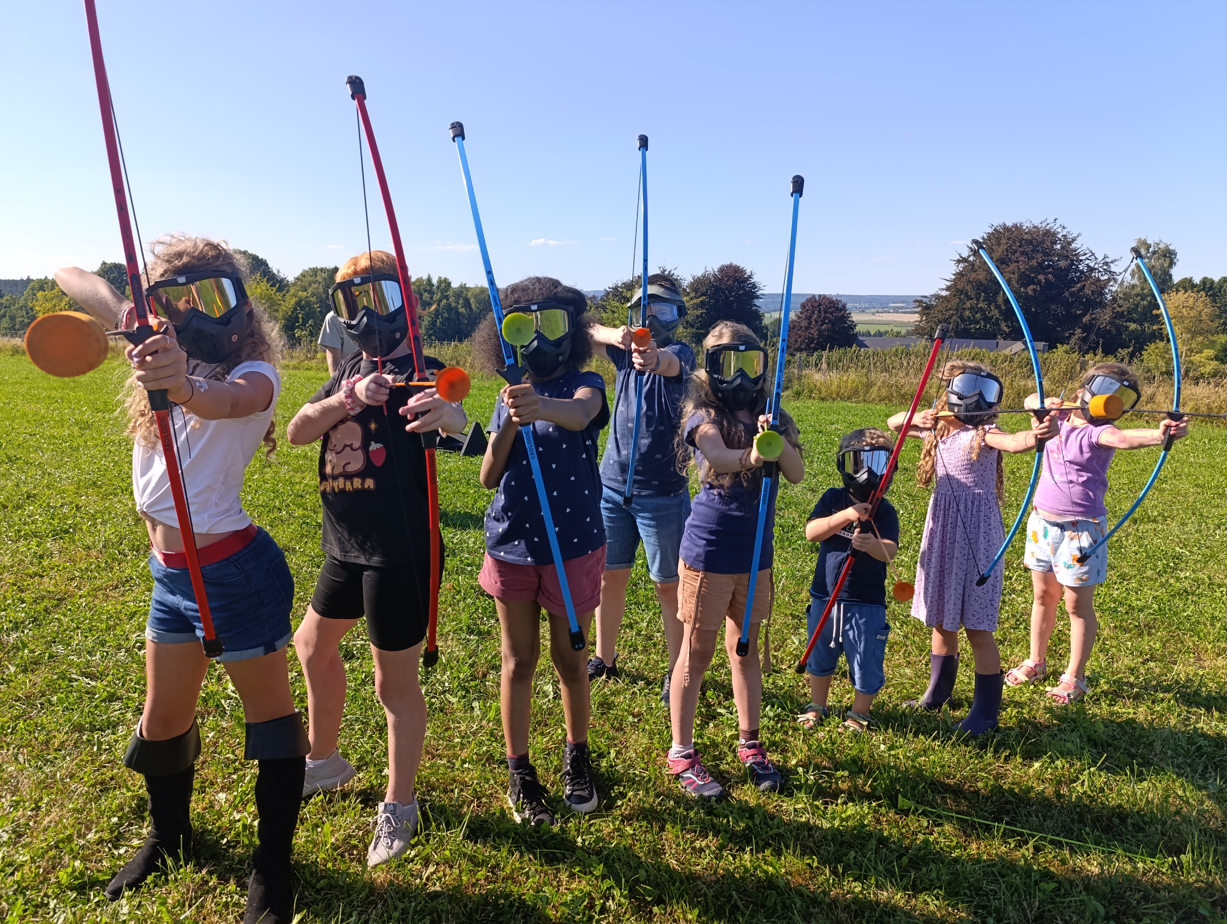 8 enfants placés côte à côte, équipés d'un masque, d'un arc et d'une flèche, photographiés en train de viser
