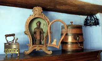Sur un buffet du milieu du XIXe siècle se trouvent trois objets : un fer à repasser à braises en fonte posé sur son support en laiton ajouré, une niche vitrée en bois sculpté contenant une statue en bois de Saint Hubert et une baratte en chêne en forme de tonnelet décorée d’un cœur en fer. Une couronne d’office ornée alternativement de fleurs de lys et d’oiseaux est en arrière-plan. Ⓒ 2024 CC BY-NC 4.0 Gilles Pirlot de Corbion