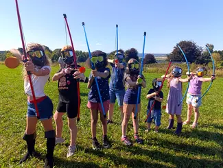 8 enfants placés côte à côte, équipés d'un masque, d'un arc et d'une flèche, photographiés en train de viser